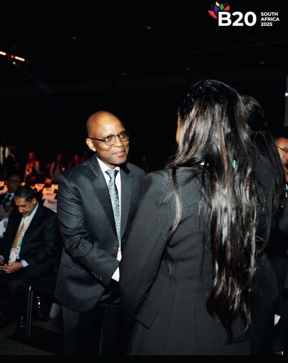 A female is backing the camera while a male is facing the camera. They're both chatting at the 2025 B20 event on the sidelines of the G-20 South Africa. They are all dressed in suits.

There's a man looking at his phone in the background alongside a group of people who are also attending the event.